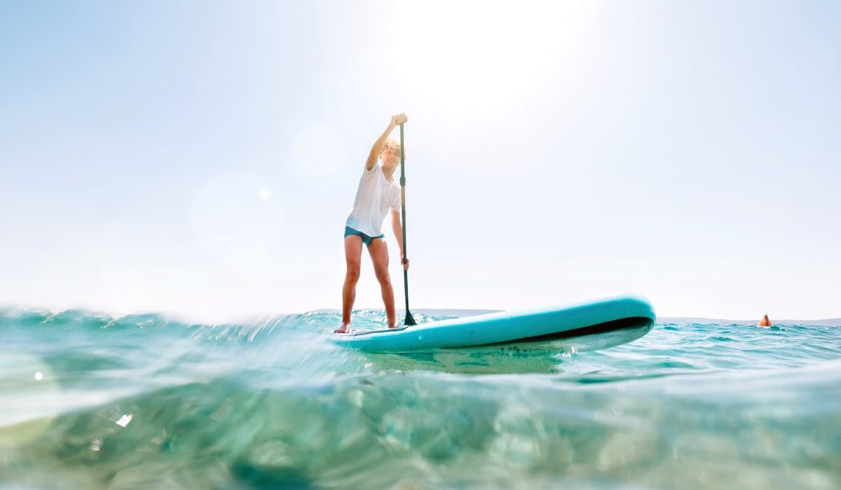 Ein Junge in Badesachen ist auf einem Stand-Up-Paddle-Board zu sehen. Er steuert das Board durch einen türkisfarbenen See.